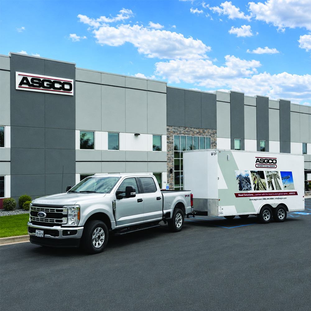 Silver pickup truck towing an ASGCO trailer in a parking lot in front of a modern industrial building with ASGCO signs on the wall.